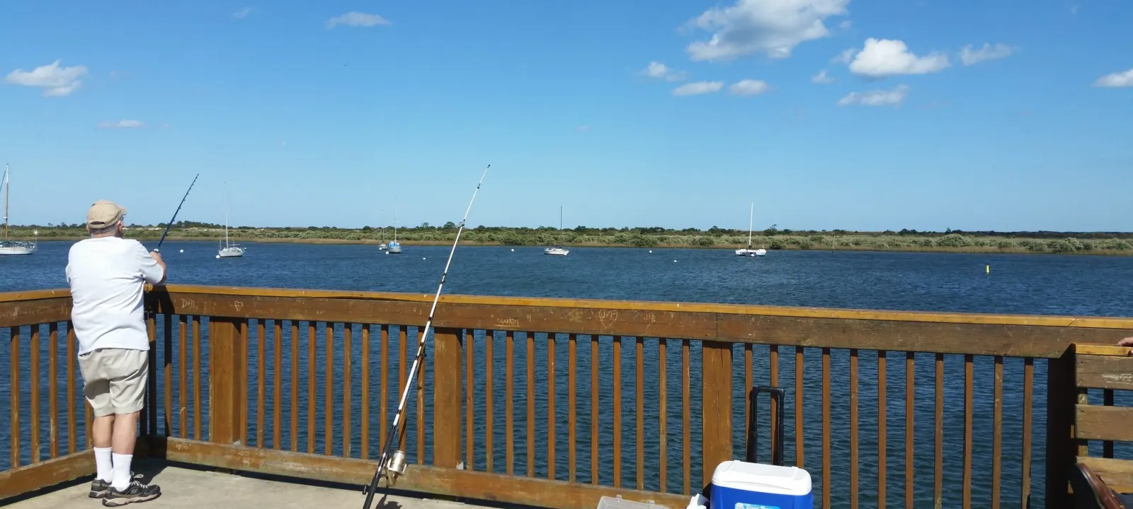 A man fishing from a pier railing with a cooler beside him