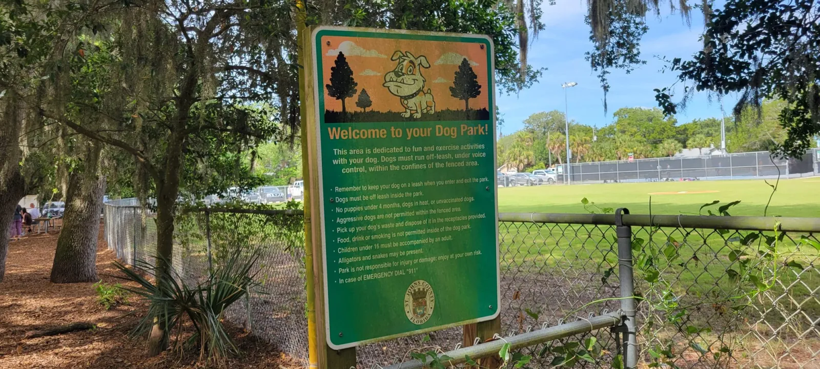 A county dog park rules sign on a chain-link fence, headed Welcome to your Dog Park! with a cartoon bulldog, under live oaks hung with Spanish moss.
