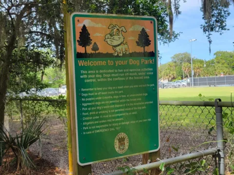 A county dog park rules sign on a chain-link fence, headed Welcome to your Dog Park! with a cartoon bulldog, under live oaks hung with Spanish moss.