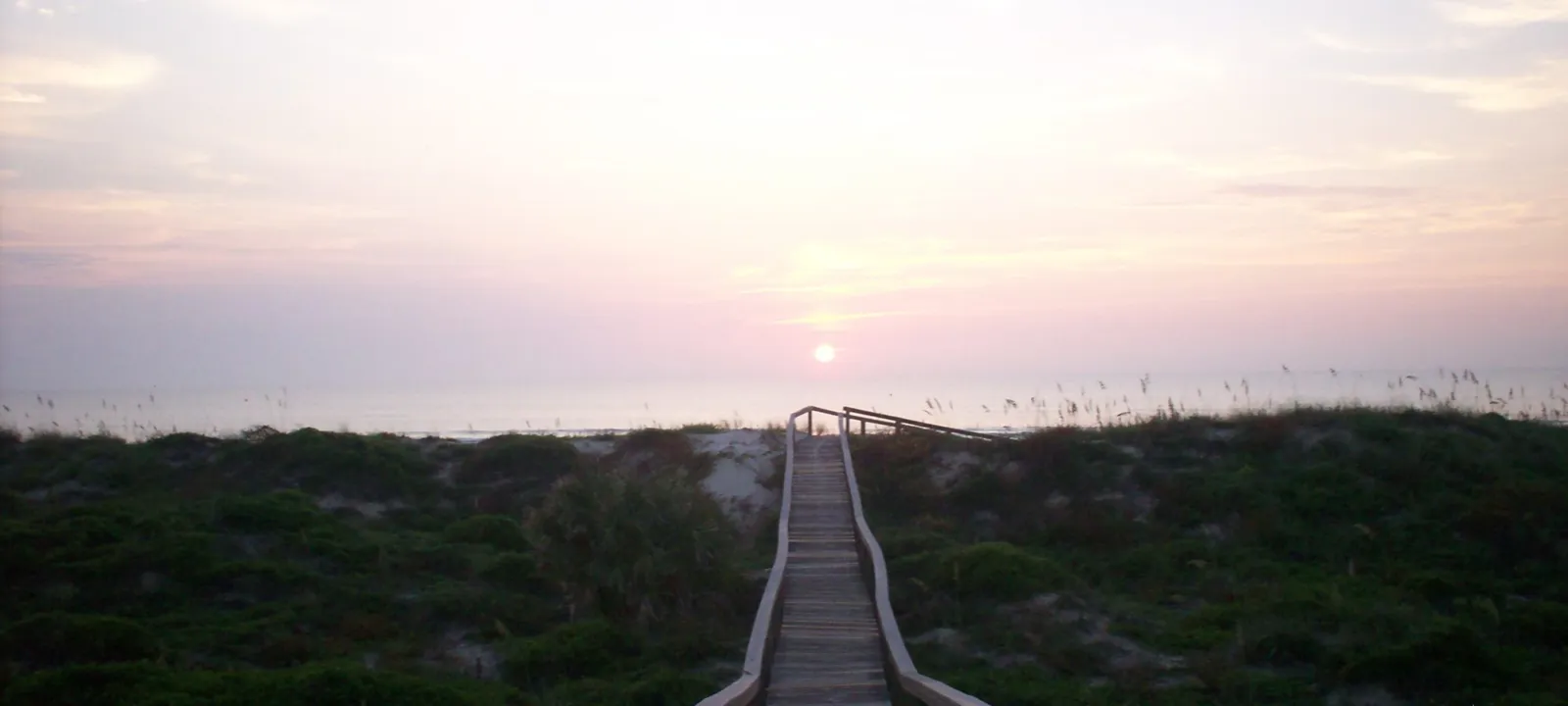 A wooden boardwalk crossing the dunes to the beach at sunrise.