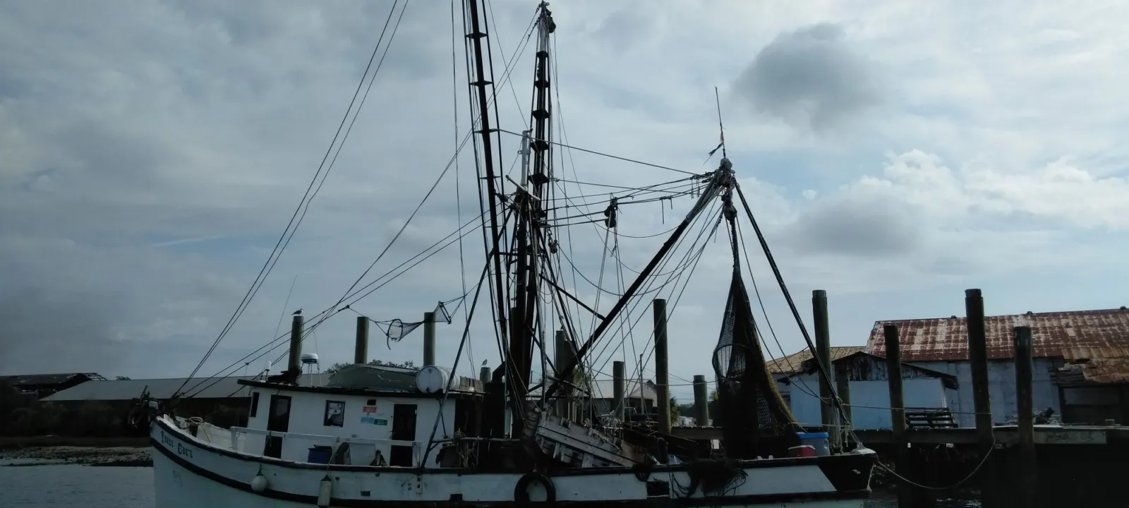 A white shrimp boat with tall rigging and hanging nets moored beside a tin-roofed dock building at Fernandina Beach.